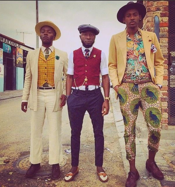 Three stylish African men wearing patterned trousers, vests and hats in urban street setting