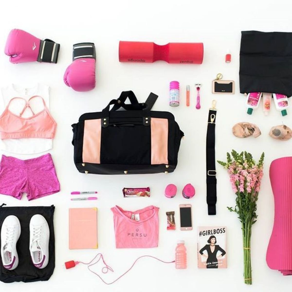 Flat lay of women’s gym essentials arranged around a black and blush duffel bag including pink boxing gloves, sports bra, shorts, water bottle, sneakers, yoga mat, fitness tracker, notebook, and flowers